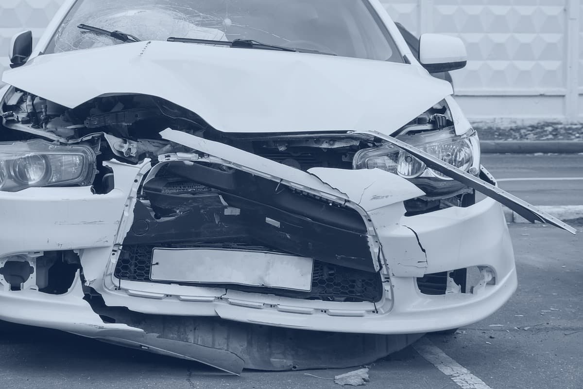 Front end of a white sedan after a collision, hood buckled and the bumper cover torn away from both headlamps.