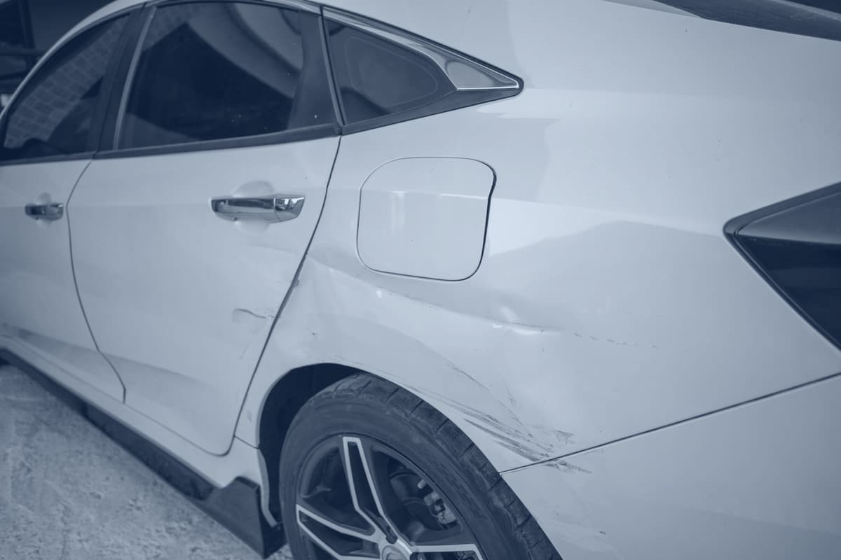Rear quarter of a white hatchback, rear door and quarter panel creased with a scrape running down to the wheel arch.